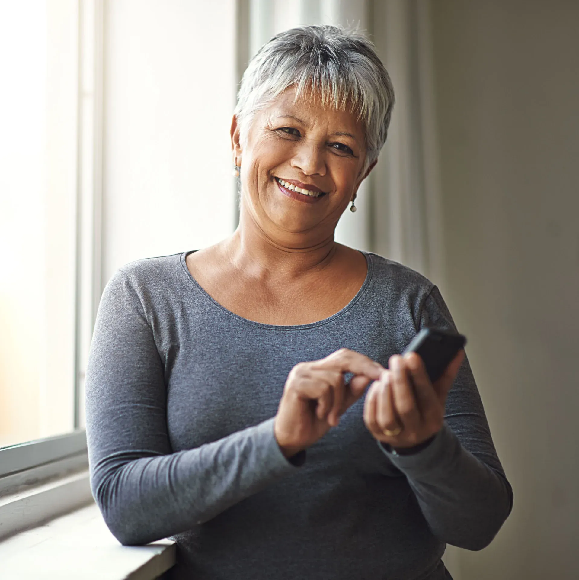Older woman smiling while using her phone by a window